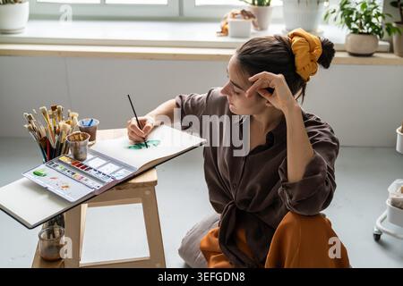 Artista donna di talento che trascorre del tempo libero con hobby d'arte, dipingendo immagini in un libro di schizzi con aquarelle, donna spensierata seduta sul pavimento disegnando in album, godendo di arte terapia e abilità formative. Foto Stock
