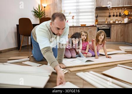 Un padre guida i suoi tre figli in un progetto fai-da-te, assemblando mobili su un pavimento di legno nel loro caldo e invitante salotto durante un pomeriggio di sole. Foto Stock