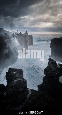 Vista aerea di una persona in piedi su scogliere vulcaniche con le braccia sollevate mentre le onde si schiantano contro le rocce scure sotto un cielo spettacolare a la Reunion, Nouvell Foto Stock