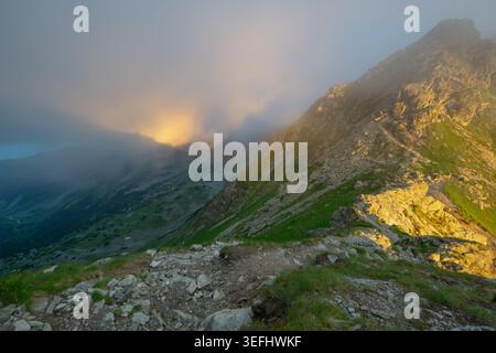 Vista del berlo Smutne nei Tatra occidentali con luce del sole dorata che illumina la cresta rocciosa e la fitta nebbia che si snoda sui verdi pendii della montagna Ziar, Zi Foto Stock