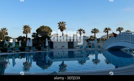 Area piscina esterna con palme e sedie a sdraio, che riflettono il cielo blu e le nuvole, caratterizzata da un ponte bianco e da una moderna struttura a bordo piscina Foto Stock
