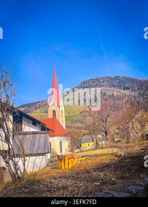 Chiesa del villaggio in piedi su una collina a Luson, in Italia, circondata da case tradizionali, alberi e montagne verdi ondulate Foto Stock