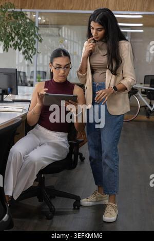 Diverse collaboratrici di sesso femminile in Borgogna e beige che collaborano su tablet in ufficio vicino alla bicicletta Foto Stock