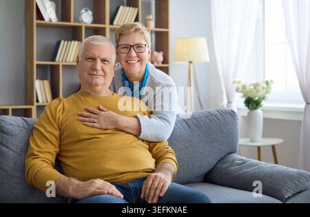 Felice coppia di anziani che si abbraccia a casa sul divano, allegro ritratto in un accogliente soggiorno Foto Stock