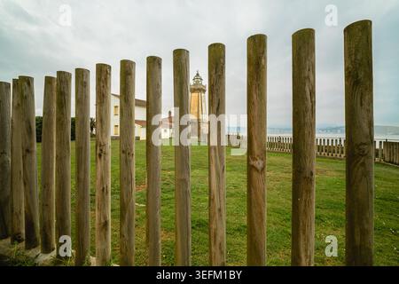 I pali di legno incorniciano una vista costiera dello storico faro di Aviles e dei custodi che costruiscono contro l'erba verde, il cielo nuvoloso e il lontano sho asturiano Foto Stock