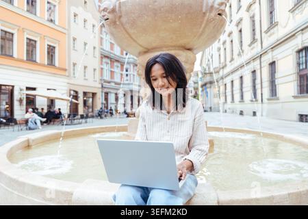 Giovane donna asiatica che lavora con un computer portatile seduto accanto a una fontana di pietra nella città europea Foto Stock