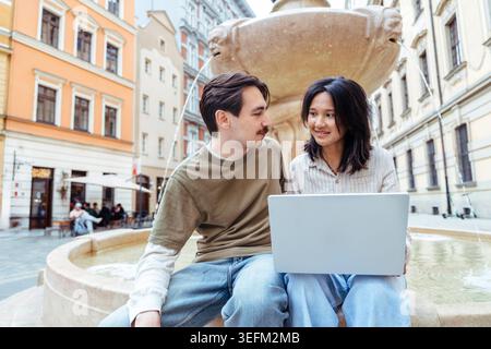 Felice giovane coppia che usa un computer portatile insieme mentre si siede accanto alla fontana nella città europea Foto Stock
