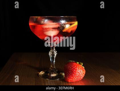 Aperitivo rosso rinfrescante con ghiaccio, agrumi e fragole fresche in un bicchiere coupé su sfondo scuro cocktail fresco ed elegante perfetto per l'estate Foto Stock
