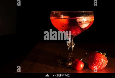 Cocktail rosso in un bicchiere di coupé con ghiaccio e ciliegie, che si illumina su un tavolo di legno soffice accanto a una fragola fresca, gettando ombre morbide nelle basse serate Foto Stock