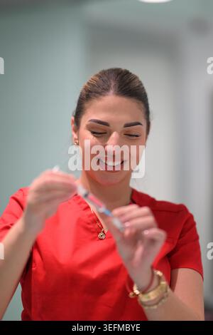 Infermiera donna sorridente che prepara la siringa vaccino in clinica che indossa scrub rossi e gioielli d'oro Foto Stock