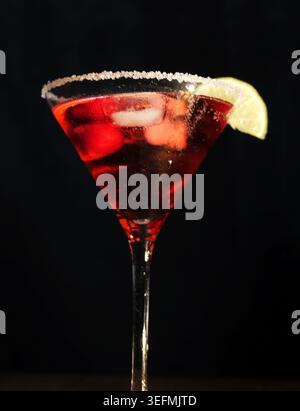 Cocktail rosso cosmopolita servito in un bicchiere di martini con ghiaccio, guarnito con una spiccata di lime fresca e un bordo di zucchero, isolato su uno sfondo scuro per un n Foto Stock