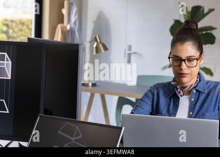 Donna che indossa camicia e sciarpa in denim e lavora su un laptop con due monitor in studio Foto Stock