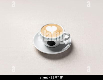 Un cappuccino con crema di latte decorato con latte art a cuore servito in una tazza bianca su un piattino. Le tonalità calde e la luce soffusa lo rendono ideale per un caffè Foto Stock