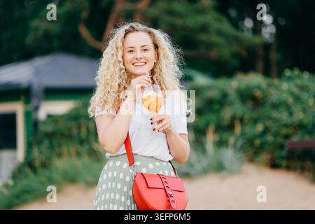 Donna europea felice con capelli biondi ricci, indossa abiti casual, tiene una borsa rossa elegante, beve un cocktail estivo fresco, cammina nel parco, sembra gioiosamente in lontananza si trova all'aperto. Concetto di svago Foto Stock