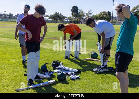Diversi compagni di squadra maschili si preparano per la pratica di cricket sul campo erboso in abbigliamento sportivo con pipistrelli. Outdoor, atleti, lavoro di squadra, allenamento, equipaggiamento, cuscinetti, casco Foto Stock