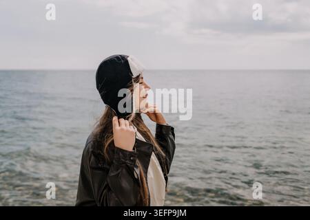 Donna ushanka Coast giovane donna che indossa un cappello invernale e una giacca in pelle guarda al mare in una giornata nuvolosa Foto Stock