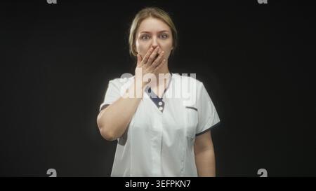Infermiera donna in uniforme bianca copre la bocca con la mano in studio, gli occhi spalancati e la bocca aperta visibile; sorpresa. Foto Stock