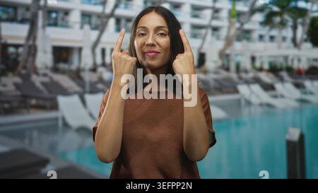 Donna ispanica punta entrambe le dita indice verso l'alto sui lettini della piscina all'aperto dell'hotel mentre sorride; fiducia giocosa. Foto Stock