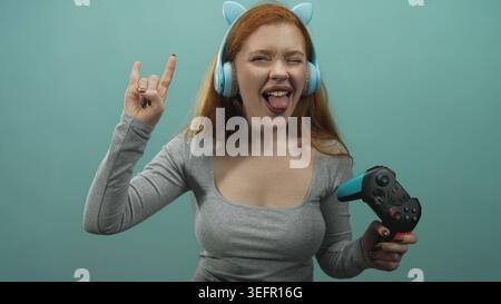Donna rossa che indossa le cuffie con orecchie di gatto e tiene in mano un gamepad, che fa lampeggiare le corna rock con la mano e la lingua in studio; giocosa. Foto Stock