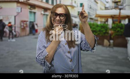 Donna anziana che sorride, tiene gli occhiali e punta il dito su una strada pedonale vicino a ombrelloni e piantagioni vicino a edifici pastello; disco gioioso Foto Stock