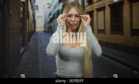 Donna che tiene gli occhiali da vista e sorride su una stretta strada acciottolata tra vecchi edifici, capelli lunghi visibili e scollatura; sicurezza. Foto Stock