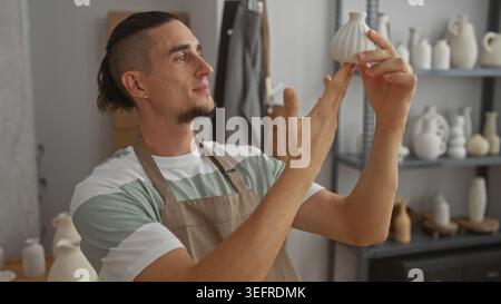 Uomo in grembiule che tiene con entrambe le mani un piccolo vaso di ceramica bianca e ne ispeziona la forma in uno studio; concentrazione artigianale. Foto Stock