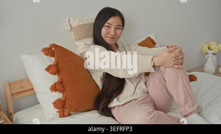 Woman sits on bedroom bed hugging knee with clasped hands and smiling beside orange tassel pillow; serenity. Foto Stock