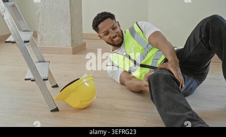 Uomo che stringe una gamba ferita accanto al cappello e alla scala caduti su un pavimento di costruzione in un edificio; dolore da incidente. Foto Stock