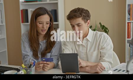Donna in possesso della carta di credito e che legge i dati mentre l'uomo tocca tablet e punti sullo schermo in un edificio per uffici; preoccupazione finanziaria. Foto Stock