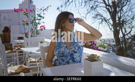 La giovane donna adatta gli occhiali da sole con la mano mentre siede a un tavolo bianco su un ristorante con terrazza dell'edificio a santorini, sorridendo verso il mare; viaggia con i leis Foto Stock