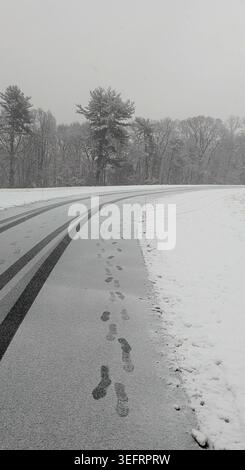 Impronte umane e tracce di pneumatici su strada asfaltata bagnata durante le nevicate invernali. Foto Stock
