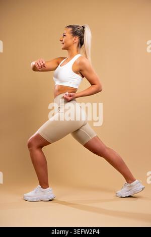 Donna atletica in reggiseno sportivo bianco e leggings beige che esegue affondi in stile studio Foto Stock