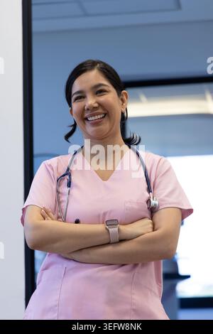Infermiera in piedi sorridente in clinica vicino alla finestra con scrub rosa, stetoscopio, smartwatch Foto Stock