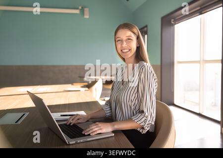 Donna caucasica che scrive sul laptop, sorride alla fotocamera in ufficio Foto Stock