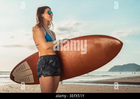Giovane donna attiva in bikini e occhiali da sole che tiene una tavola da surf su una spiaggia sabbiosa al tramonto, guardando verso l'oceano con un espresso calmo e fiducioso Foto Stock