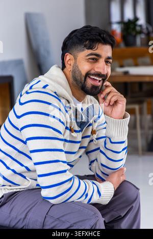 Uomo indiano seduto su una superficie bassa a casa, indossa un maglione con cappuccio e tavolo di legno dietro Foto Stock