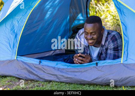 Uomo afro-americano disteso in tenda blu sull'erba, indossa una giacca a plaid con cappuccio, tiene il telefono Foto Stock
