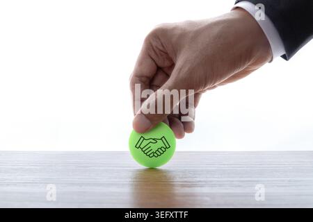Primo piano della mano di un uomo d'affari che posiziona un gettone verde con un'icona di stretta di mano su una scrivania di legno su uno sfondo bianco. Fiducia aziendale, partnership Foto Stock