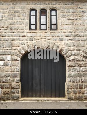 porta ad arco in legno nero su rustico muro in pietra grigia, ingresso simmetrico all'edificio storico con strette finestre, vista laterale del castello medievale Foto Stock