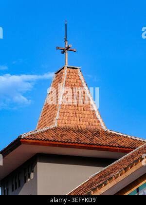 una vista dettagliata di un campanile sul tetto della chiesa con piastrelle di argilla e una croce di ferro che raggiunge un cielo blu limpido, catturato in una giornata di sole a yogyakart Foto Stock