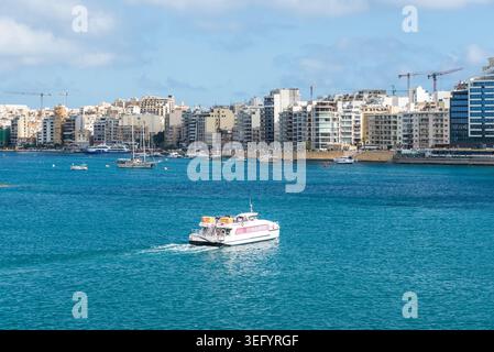 Sliema, Malta - 16 maggio 2025: Traghetto passeggeri che viaggia da Sliema a la Valletta attraverso le acque turchesi del porto di Marsamxett con il moderno specchio d'acqua Foto Stock
