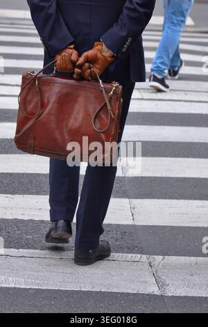 L'uomo vestito con una tuta scura e guanti in pelle marrone tiene una valigetta in pelle marrone mentre cammina attraverso un passaggio pedonale in un ambiente urbano Foto Stock