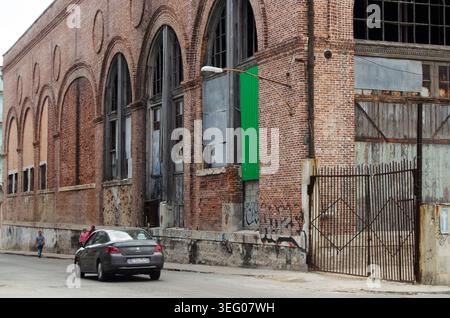 Edificio in mattoni fatiscenti con grandi finestre ad arco e un'auto che passa su una strada della città Foto Stock