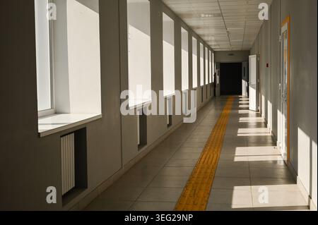 Corridoio scolastico illuminato dal sole con grandi finestre e percorso tattile giallo su pavimento piastrellato, corridoio con porte e interni luminosi, spazio didattico vuoto Foto Stock