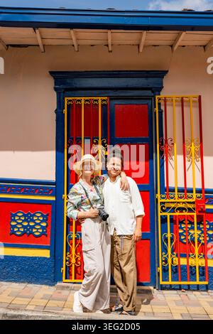 Coppia multietnica in piedi braccio di fronte a una vivace porta rossa e blu con ferro giallo a Guatape, Antioquia, Colombia, godendosi un sole Foto Stock
