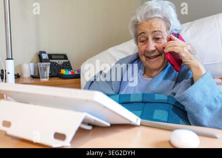 Una donna anziana in un letto d'ospedale sorride calorosamente mentre parla al telefono. Con un tablet e attrezzature mediche nelle vicinanze, appare contenuto e comfo Foto Stock