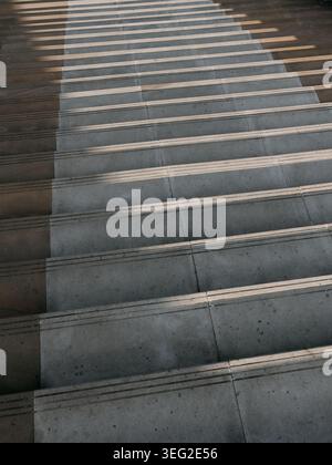 Vista astratta dei gradini delle scale in calcestruzzo con strisce antiscivolo, ombre diagonali forti e luce calda, per creare un motivo geometrico minimalista Foto Stock