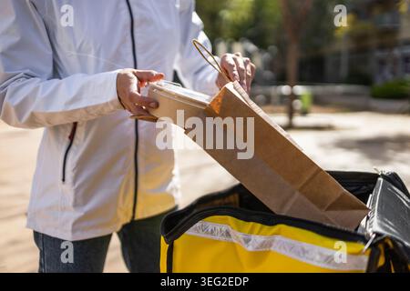 Primo piano di una donna che consegna una ciotola da asporto e un sacchetto di carta in uno zaino per corriere isolato, pronto per il servizio di consegna fast food in t Foto Stock