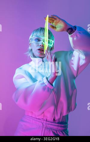 Donna futuristica, con giacca bianca e visiera al neon, regala un prisma di cristallo luminoso, che si posa sotto l'illuminazione di studio rosa e blu su uno sfondo sfumato viola Foto Stock