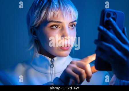Ritratto ravvicinato di una donna futuristica con capelli corti argentati che utilizza uno smartphone con una luce blu al neon, espressione mirata, atmosfera tecnologica moderna, cyber Foto Stock